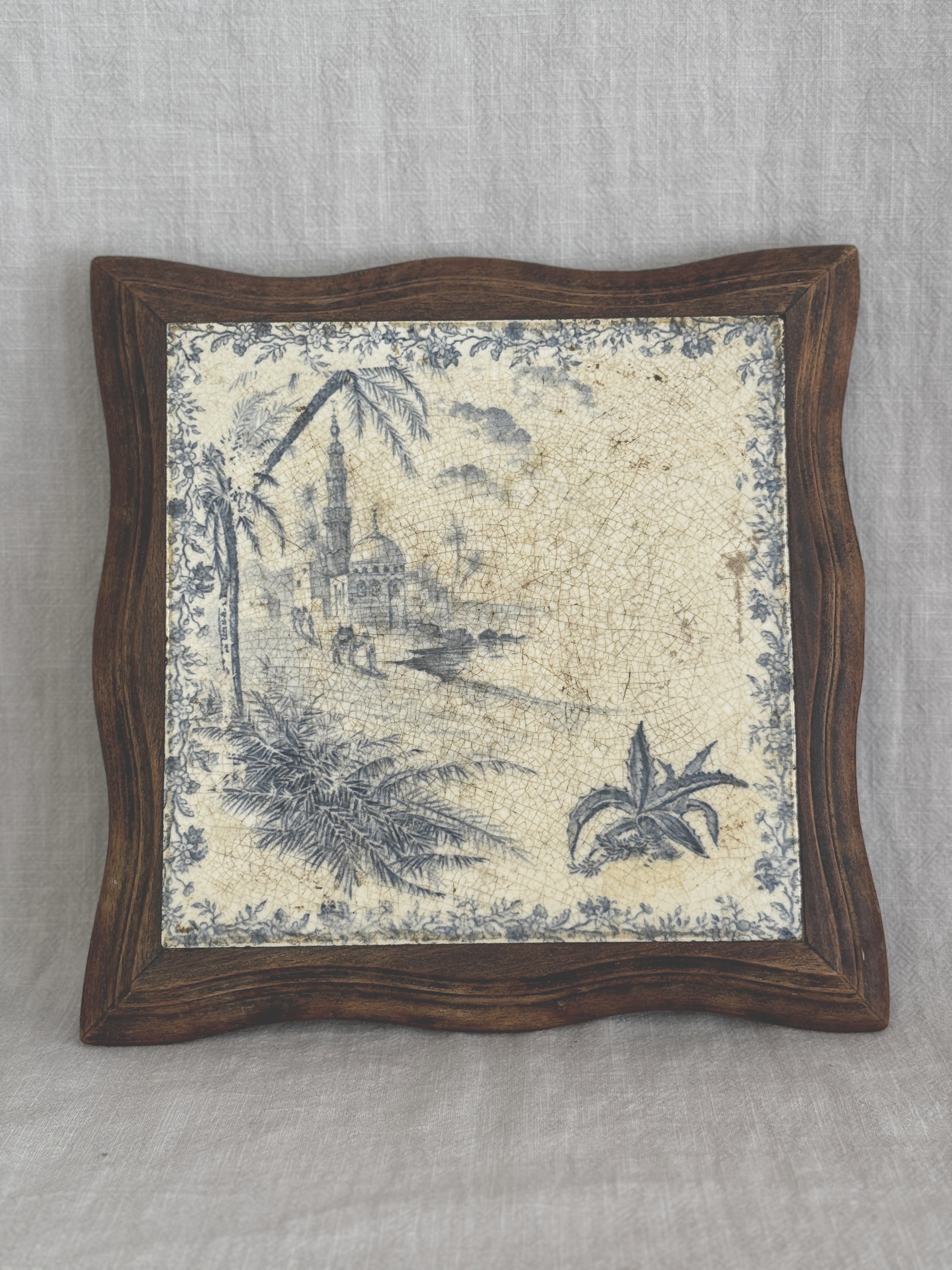 Antique French blue transferware tile trivet in a shaped wooden frame, showing a mosque, minaret and palm trees on a heavily crazed cream ground