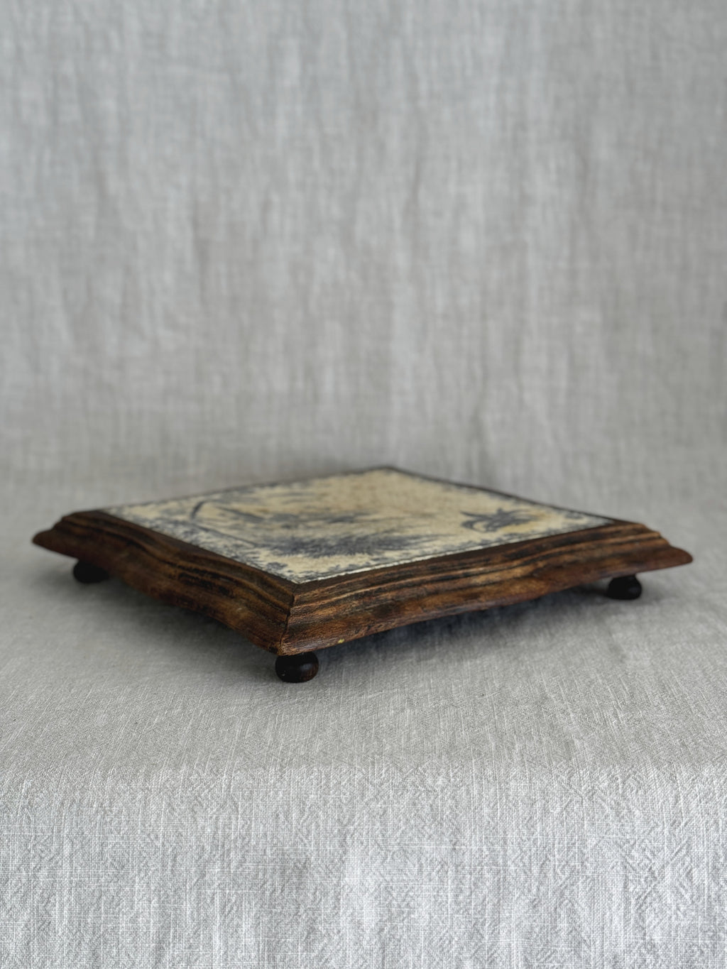 Side view of the framed French tile trivet, showing its low profile and small turned wooden feet