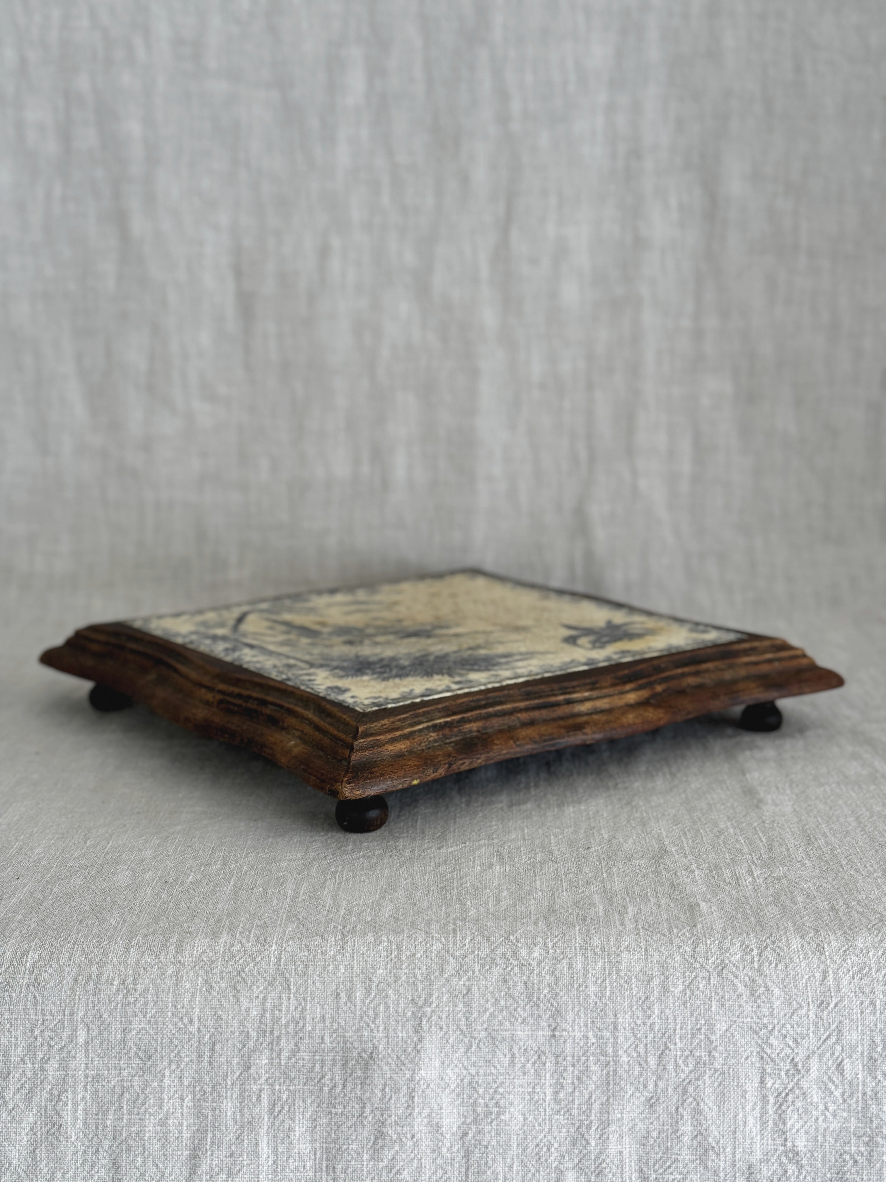 Side view of the framed French tile trivet, showing its low profile and small turned wooden feet