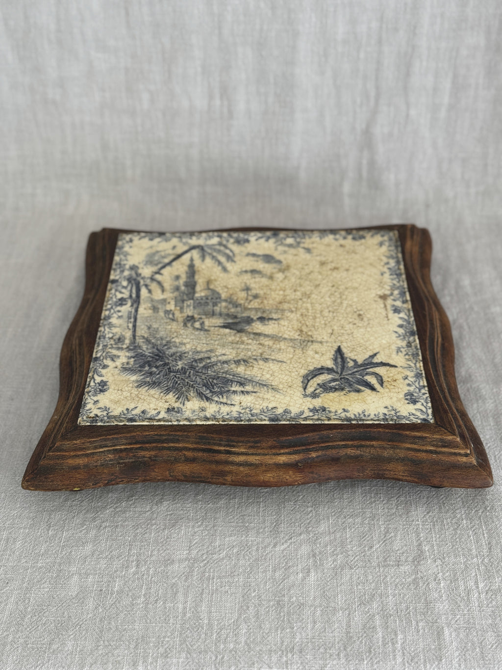 Angled view of the antique tile trivet, the blue transfer scene set into a serpentine-cut wooden frame