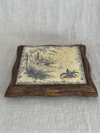 Angled view of the antique tile trivet, the blue transfer scene set into a serpentine-cut wooden frame