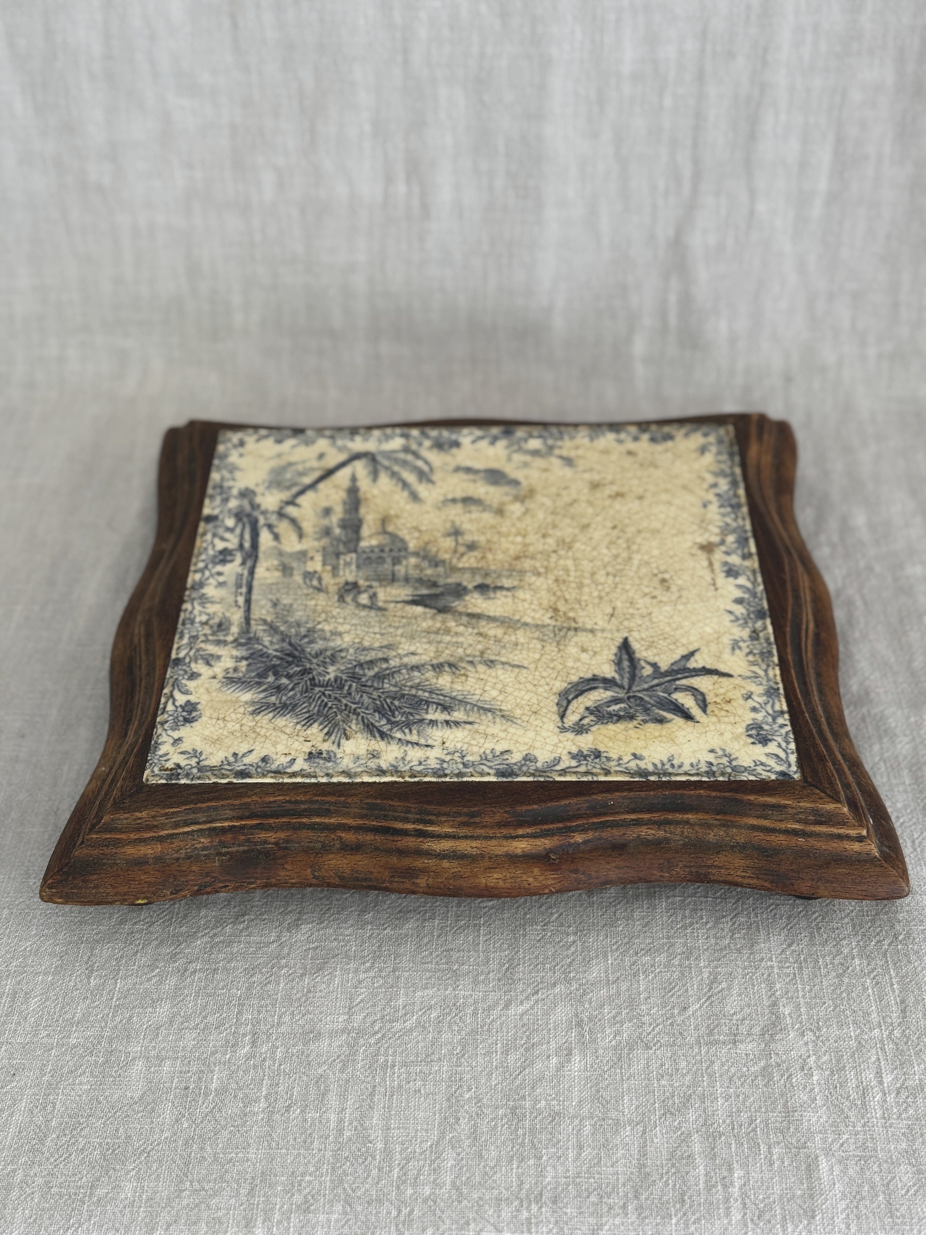 Angled view of the antique tile trivet, the blue transfer scene set into a serpentine-cut wooden frame