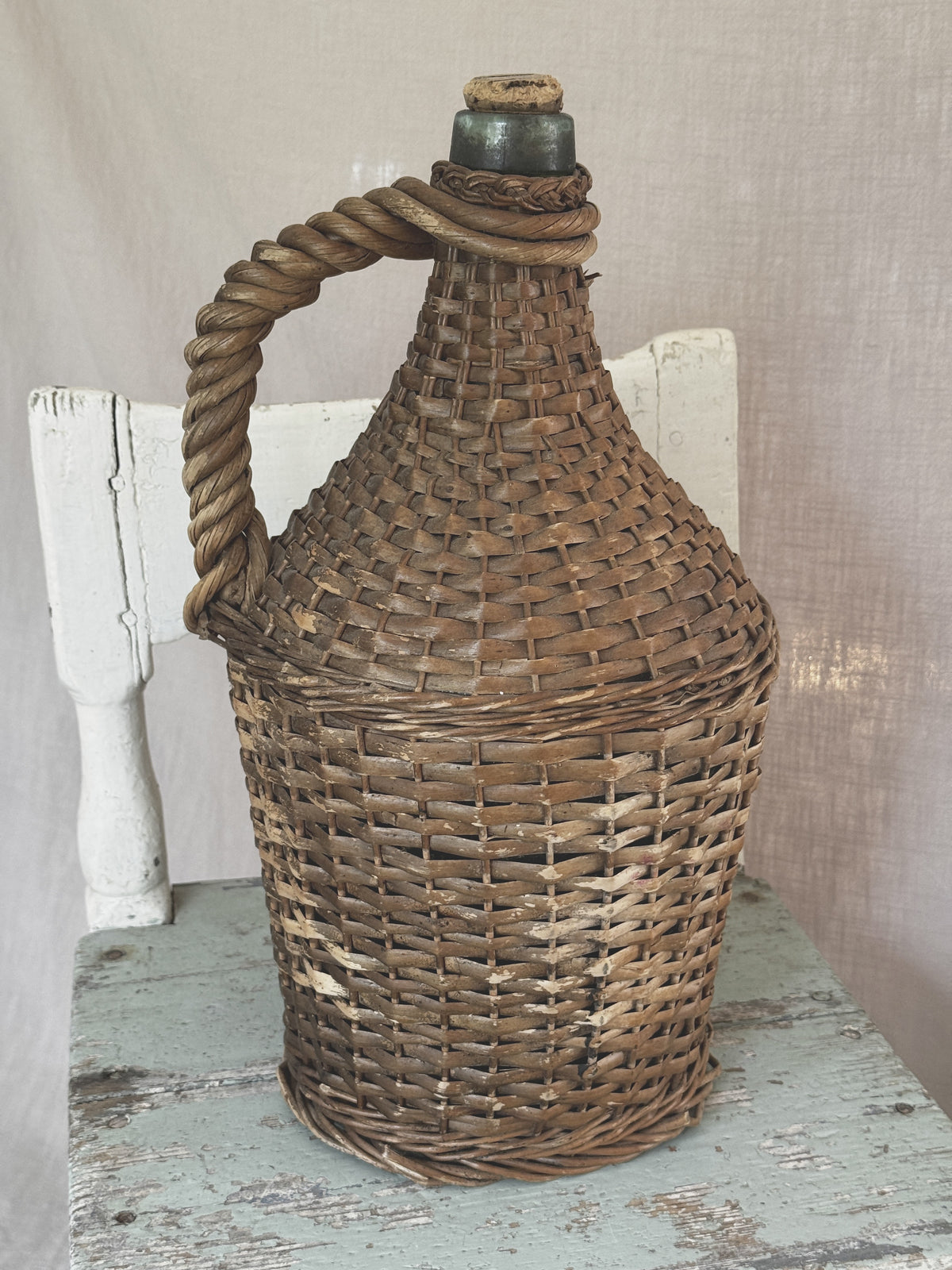 Vintage wicker demijohn in green glass with twisted rope handle and cork, standing on a painted wooden stool