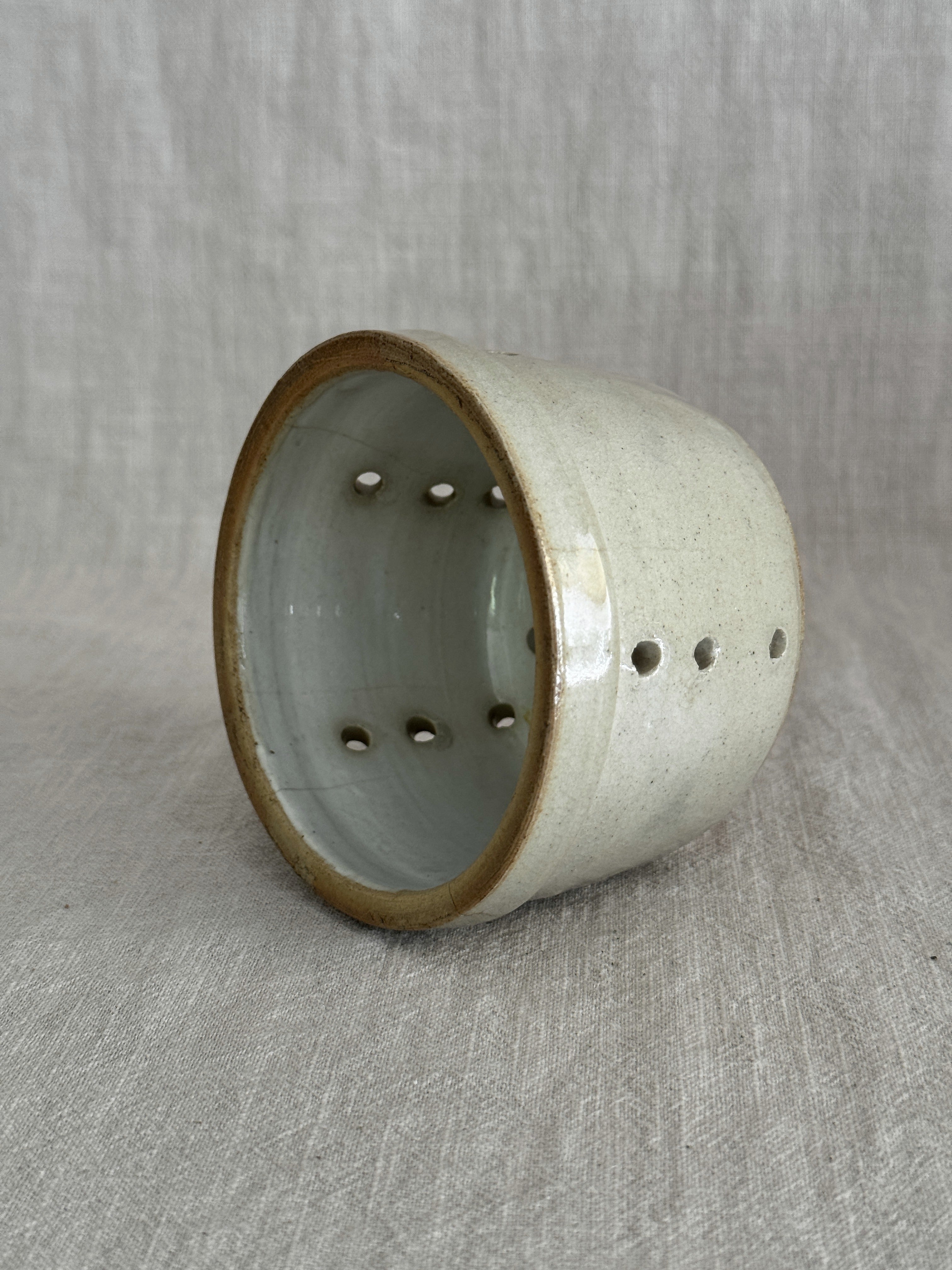 Antique French faisselle cheese mould in cream-glazed stoneware, tilted to show the drainage holes through the base