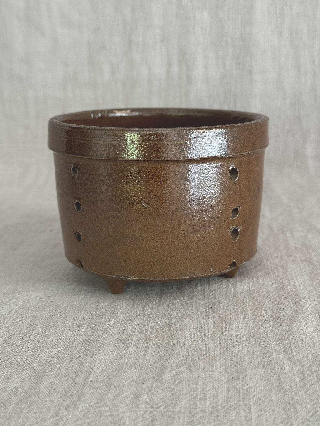 Brown salt-glazed French stoneware cheese mold standing upright on pale linen, two rows of pierced drain holes visible down the side