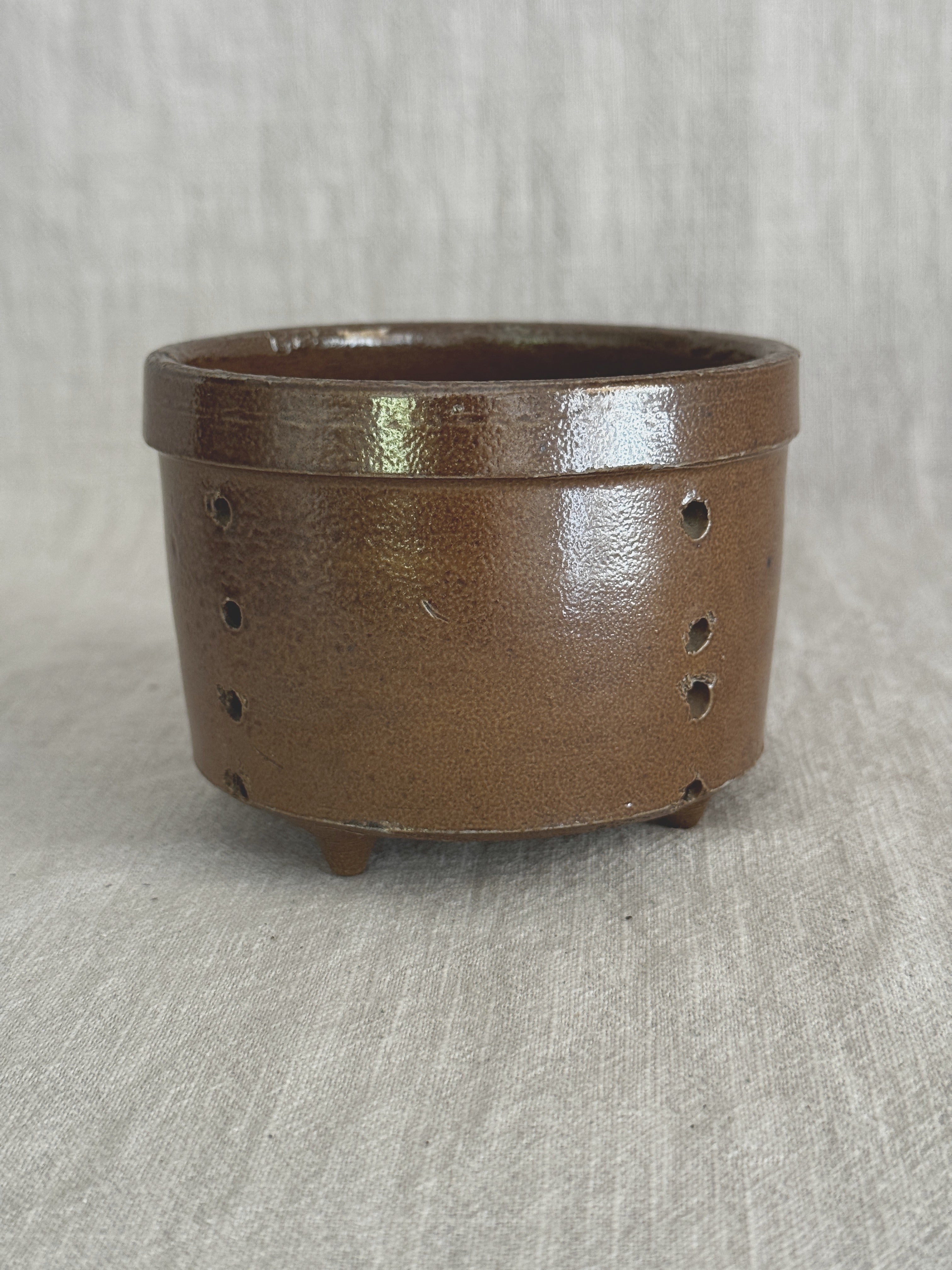 Brown salt-glazed French stoneware cheese mold standing upright on pale linen, two rows of pierced drain holes visible down the side