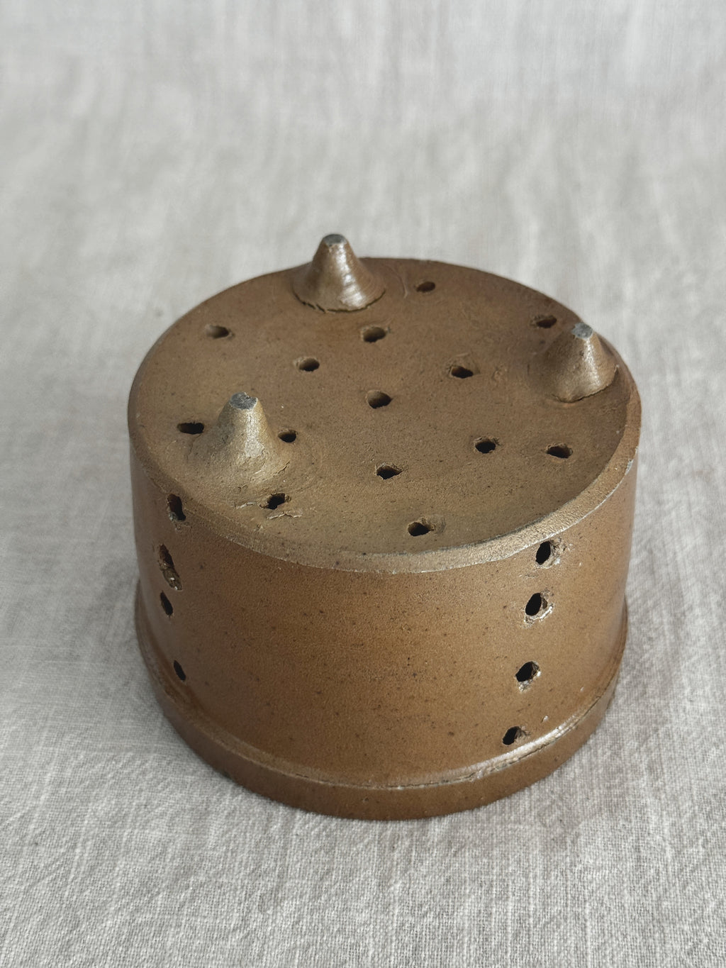 Close view of the French faisselle from below, the three clay feet and punched base holes against a linen background