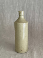Unmarked French stoneware bottle in warm putty glaze standing on linen, the long fine glaze line running down the body
