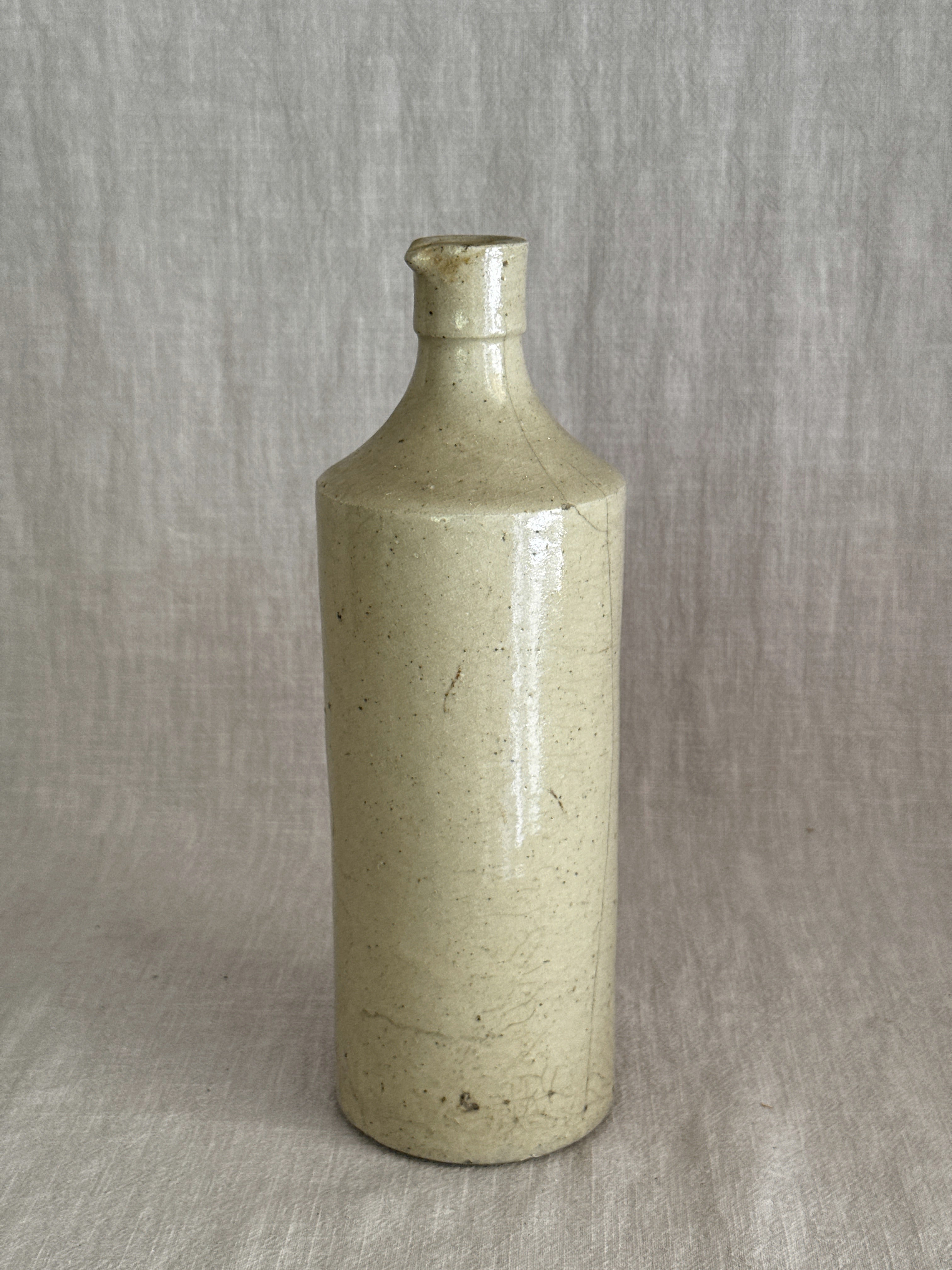 Unmarked French stoneware bottle in warm putty glaze standing on linen, the long fine glaze line running down the body