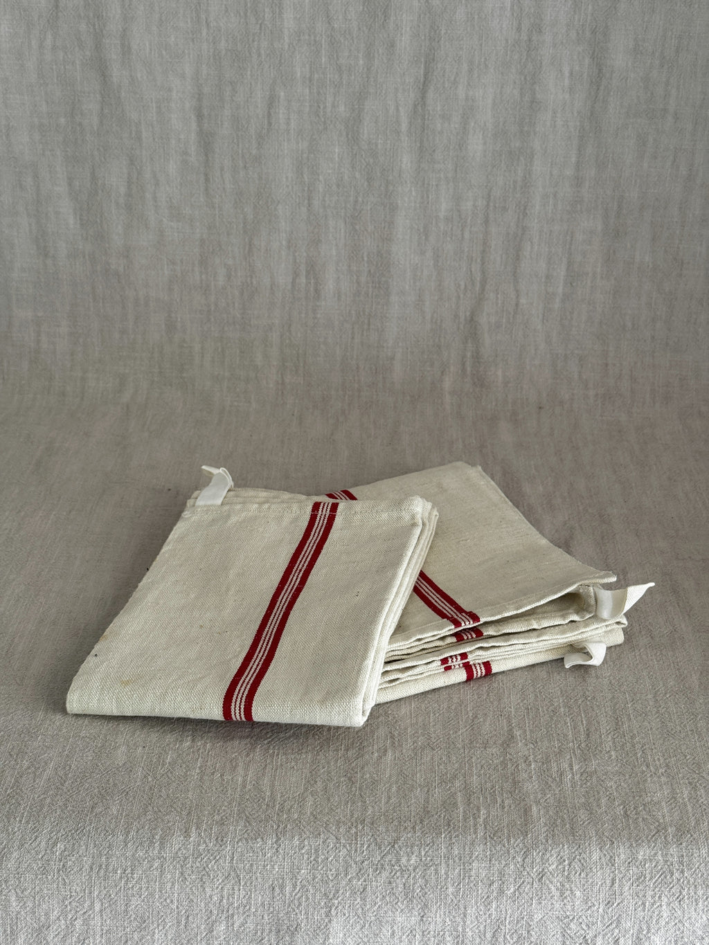 Folded cream metis linen torchon with woven red stripes and a white cotton hanging loop, resting on a linen cloth