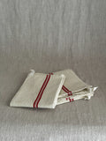 Folded cream metis linen torchon with woven red stripes and a white cotton hanging loop, resting on a linen cloth