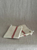 Folded cream metis linen torchon with woven red stripes and a white cotton hanging loop, resting on a linen cloth