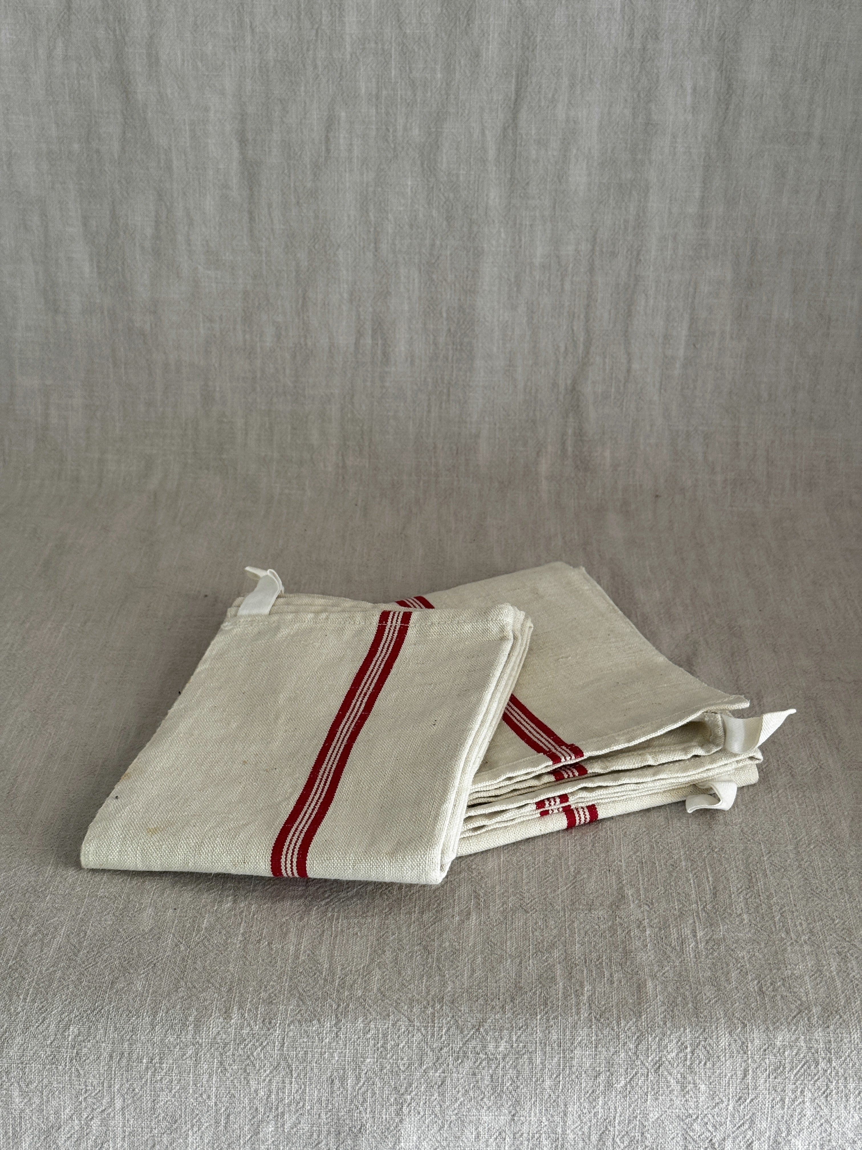 Folded cream metis linen torchon with woven red stripes and a white cotton hanging loop, resting on a linen cloth