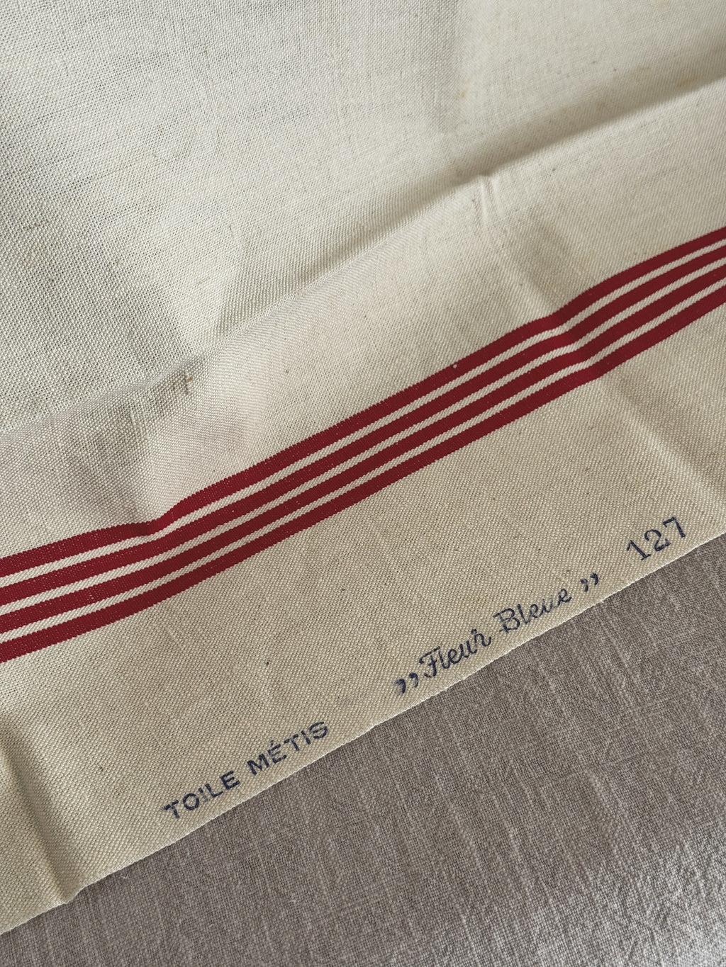 Blue printed selvedge mark reading TOILE METIS Fleur Bleue 127 beside the red woven stripes on cream metis cloth