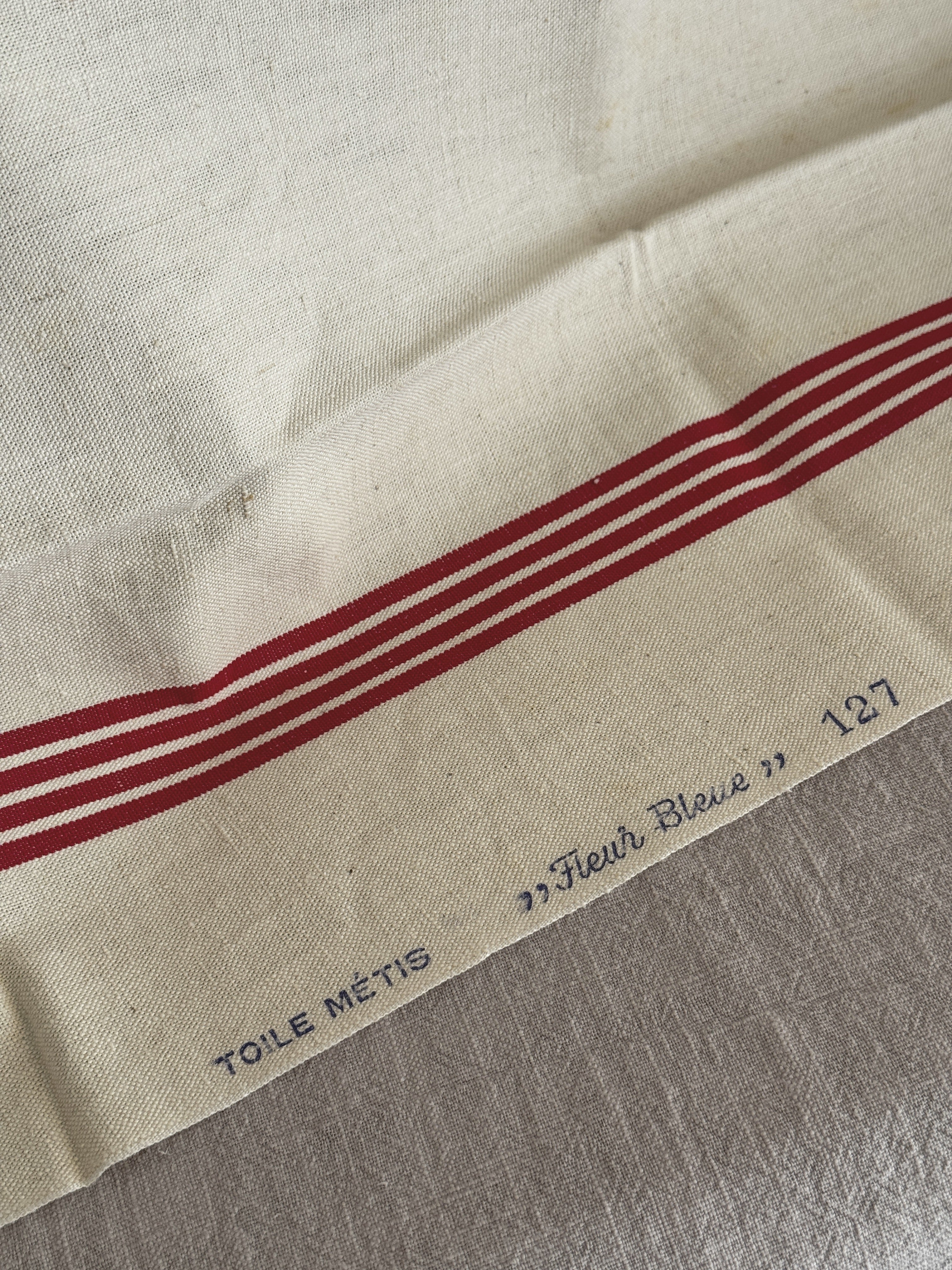 Blue printed selvedge mark reading TOILE METIS Fleur Bleue 127 beside the red woven stripes on cream metis cloth