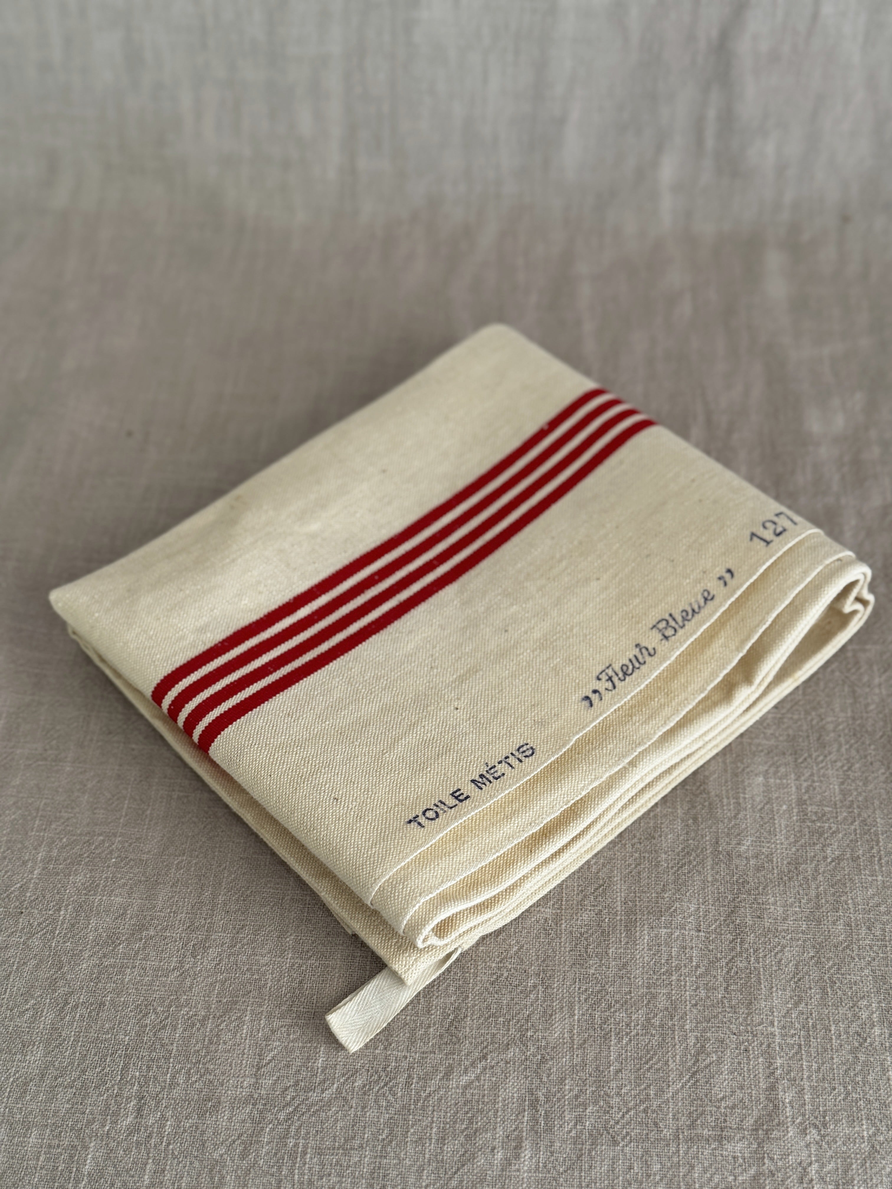 Folded unused French toile metis torchon with red stripes, cotton hanging loop and blue Fleur Bleue mark, on a grey linen surface