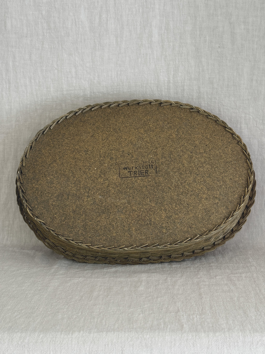 Underside of the oval willow tray showing the plain fibreboard base, the plaited edge and the maker's stamp