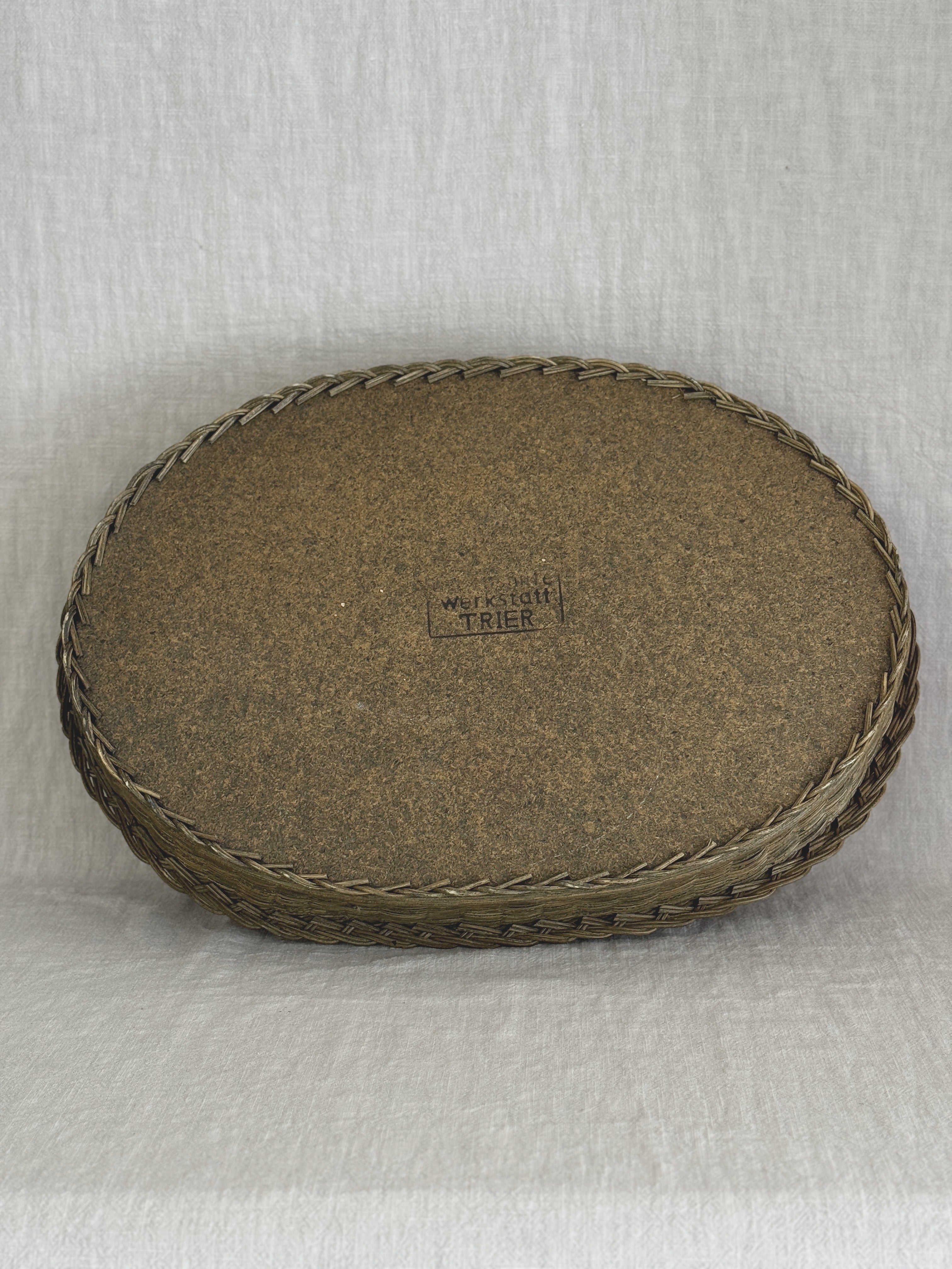 Underside of the oval willow tray showing the plain fibreboard base, the plaited edge and the maker's stamp