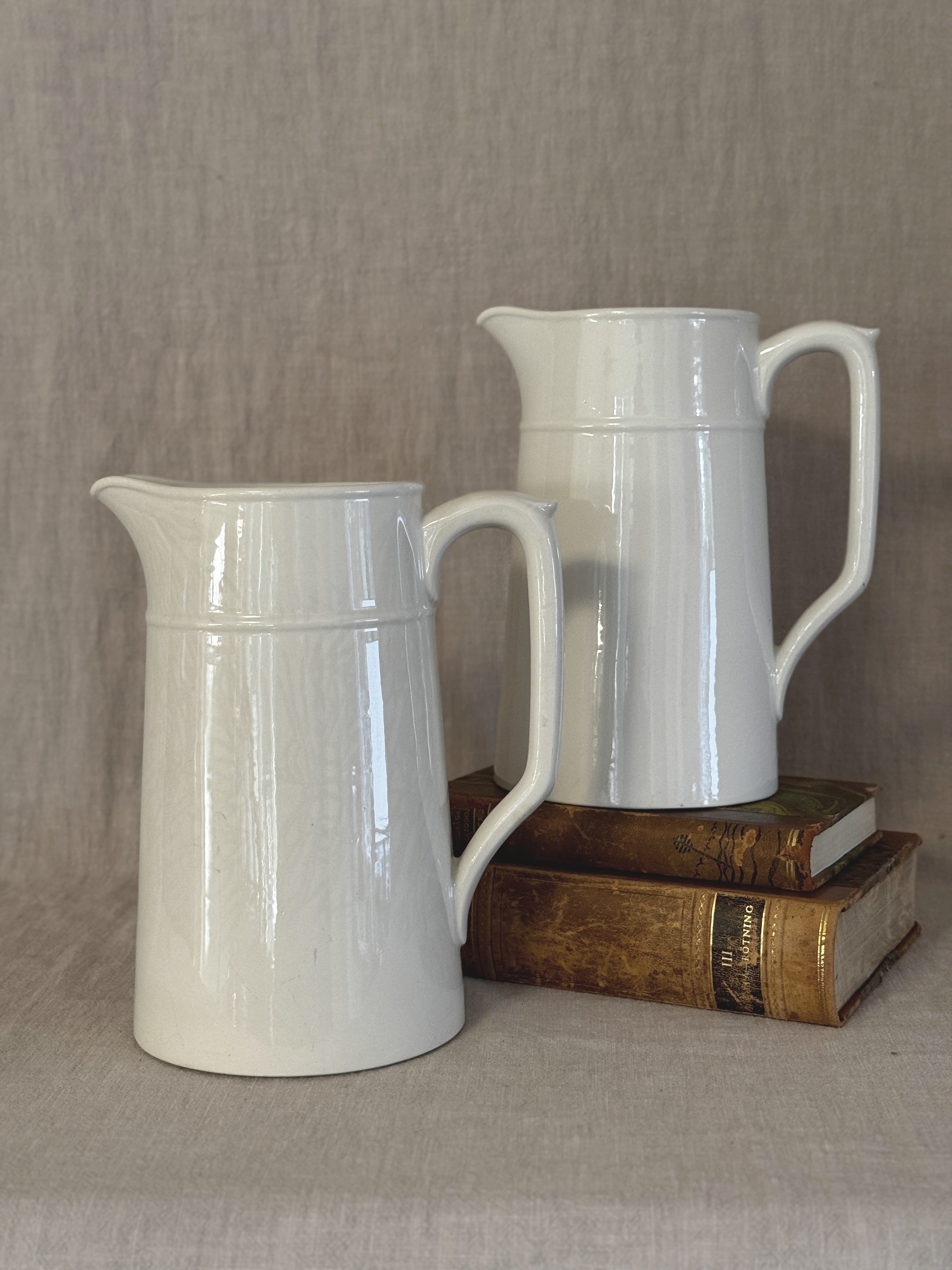 Two matching tall white Swedish jugs in plain white earthenware, styled with old books