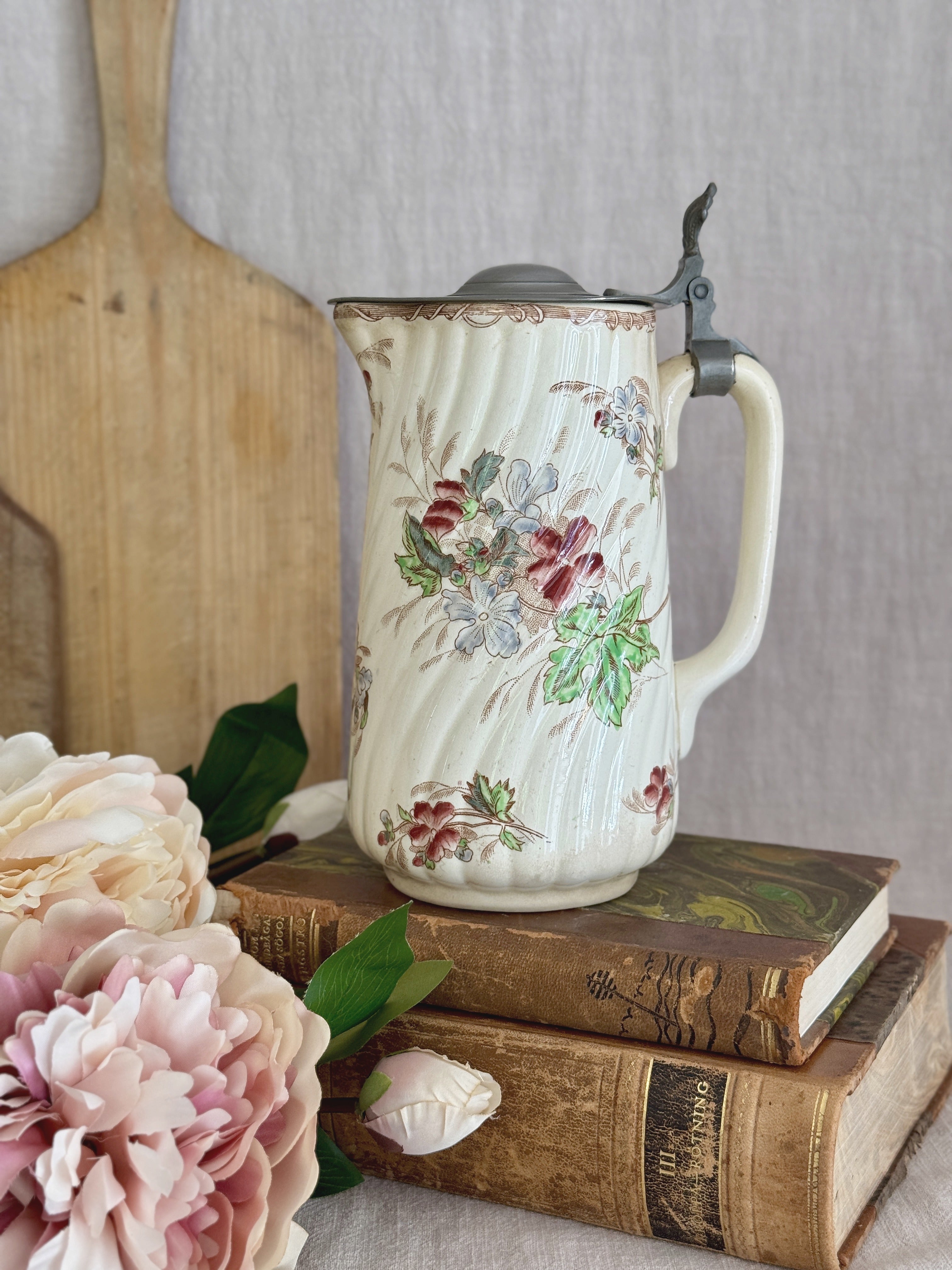 Antique French faience jug with hinged pewter lid, styled on antique books beside a wooden board and peonies