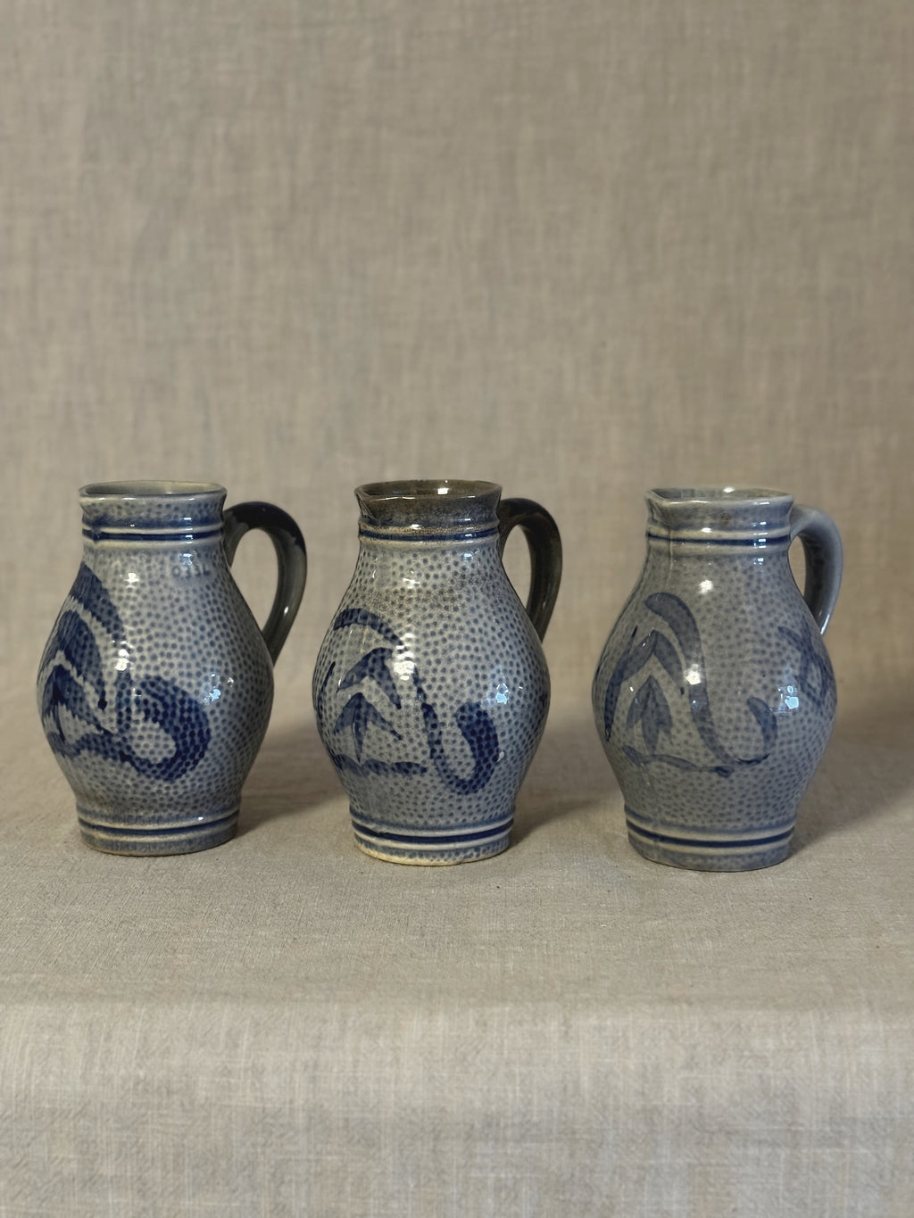 Three German salt-glazed stoneware jugs standing in a row on linen