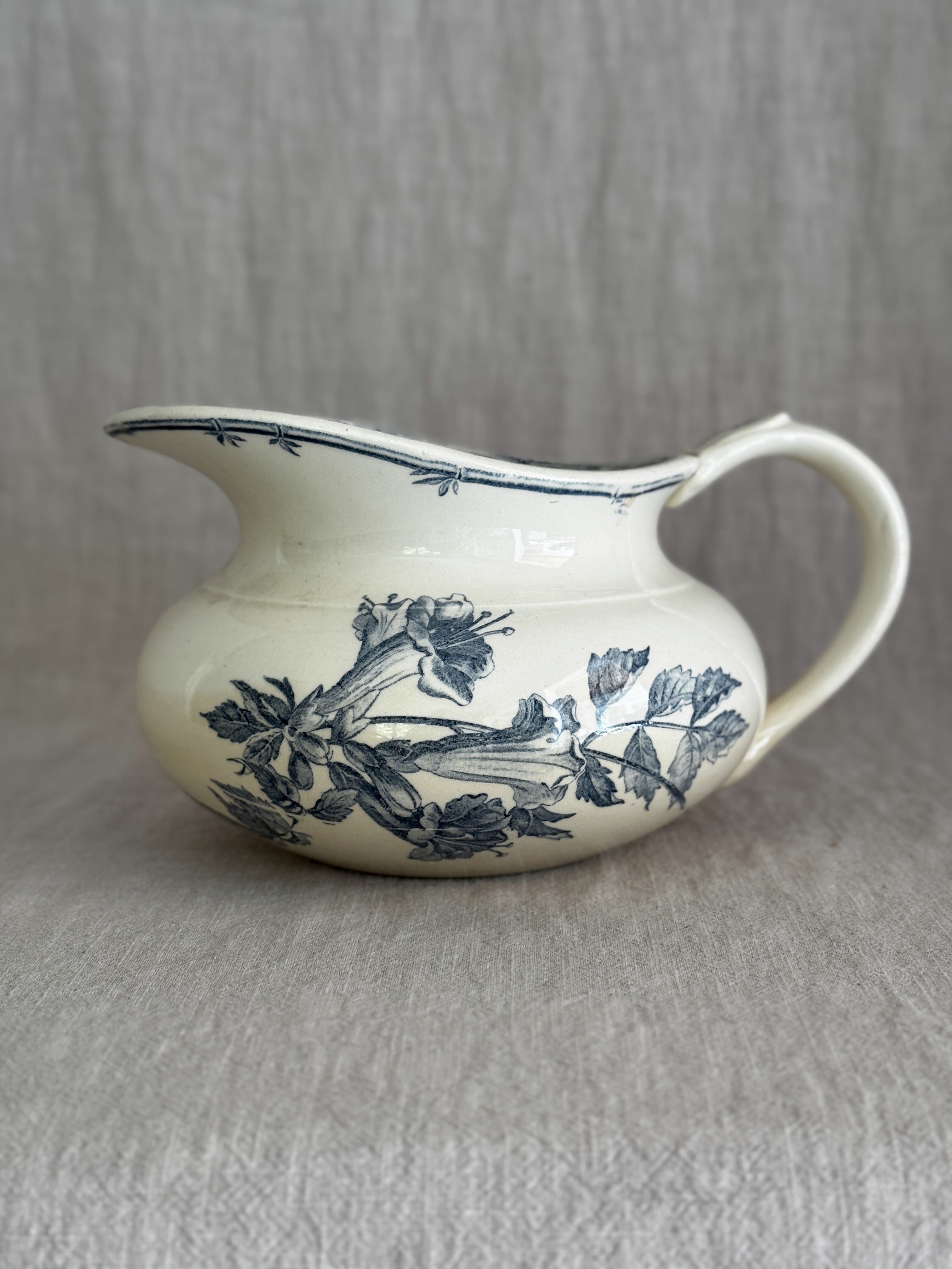 Antique Gien faience jug in cream with a blue-grey printed trumpet flower spray, a wide flat spout and a bamboo-shaped handle, on a linen backdrop