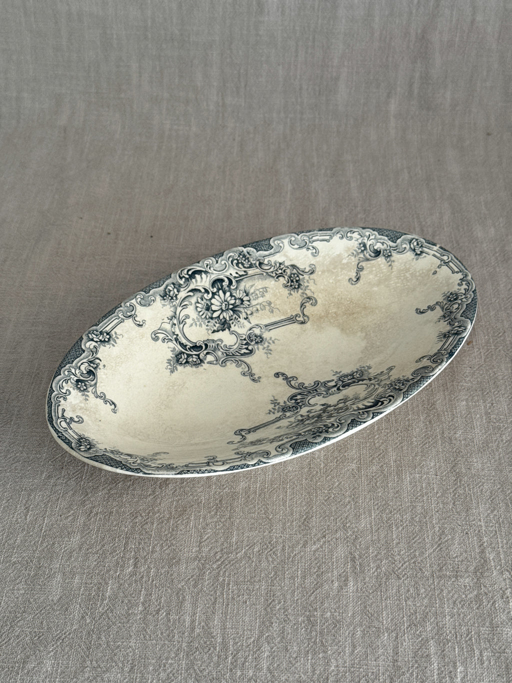 Antique Belgian oval faience ravier dish with deep teal rococo transfer print and warm tea staining, on a linen cloth