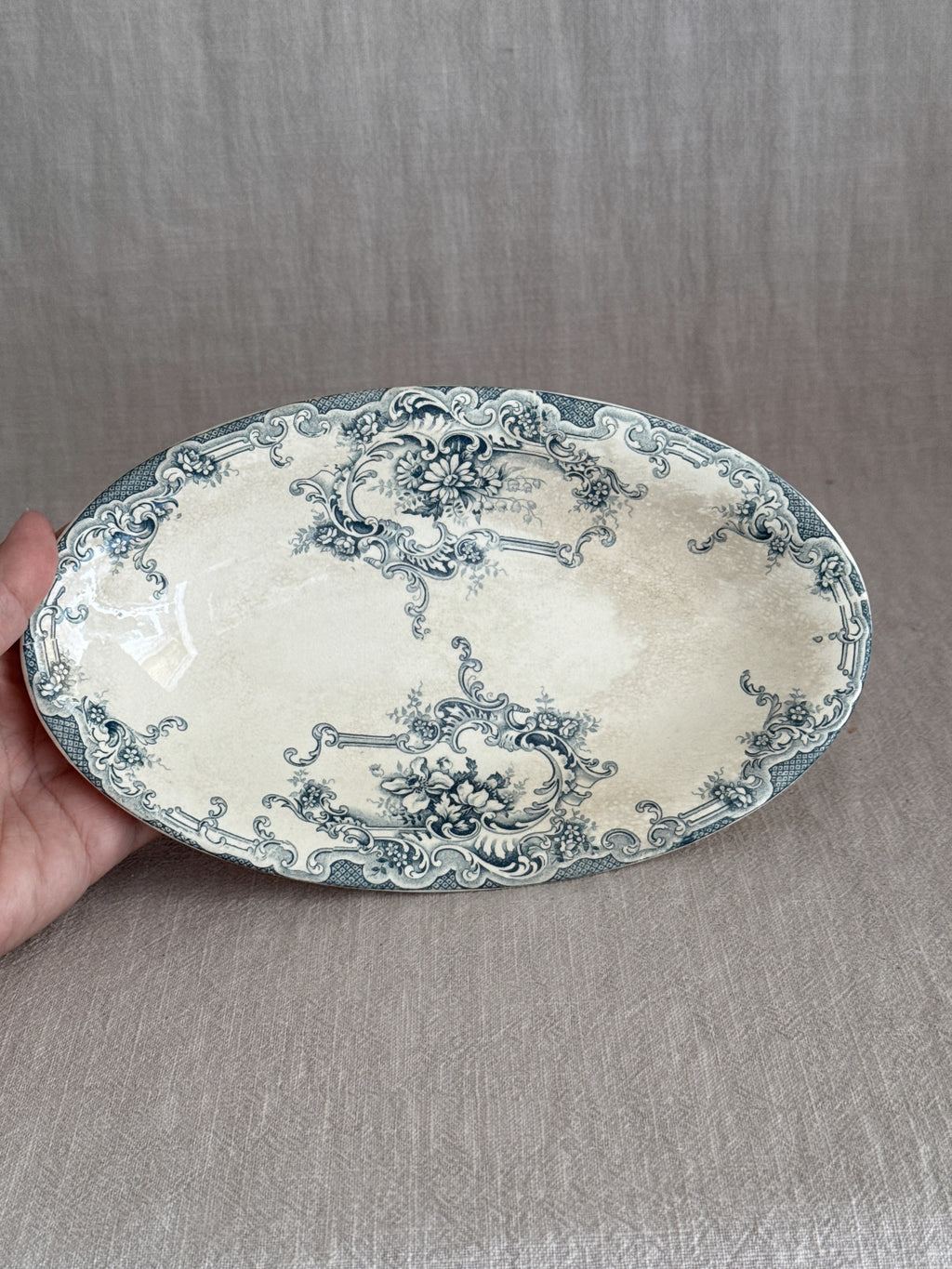 The oval faience dish held in one hand, showing the full teal transfer design of cartouches, daisies and roses