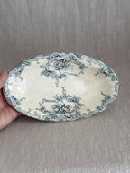 The oval faience dish held in one hand, showing the full teal transfer design of cartouches, daisies and roses
