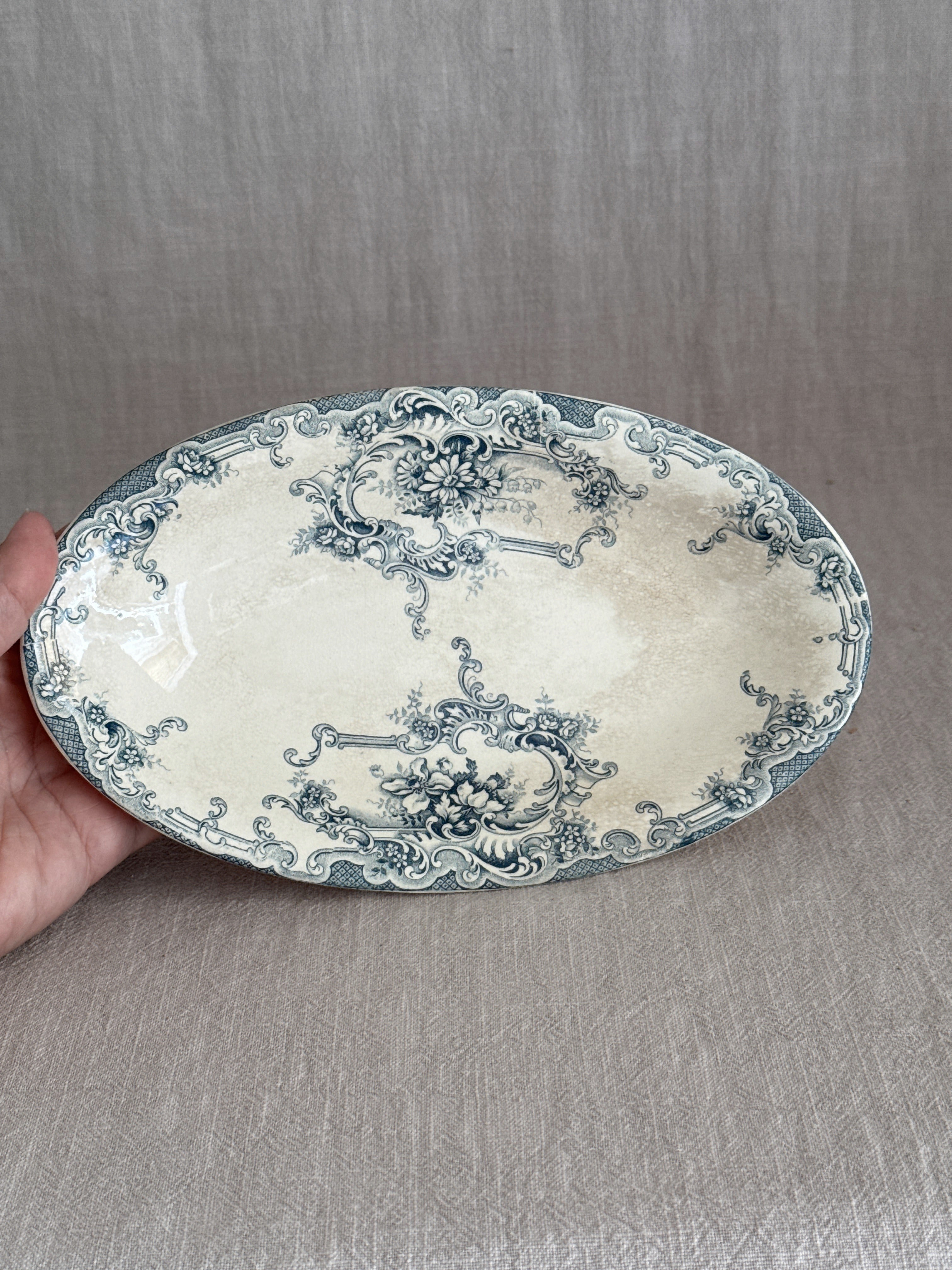 The oval faience dish held in one hand, showing the full teal transfer design of cartouches, daisies and roses