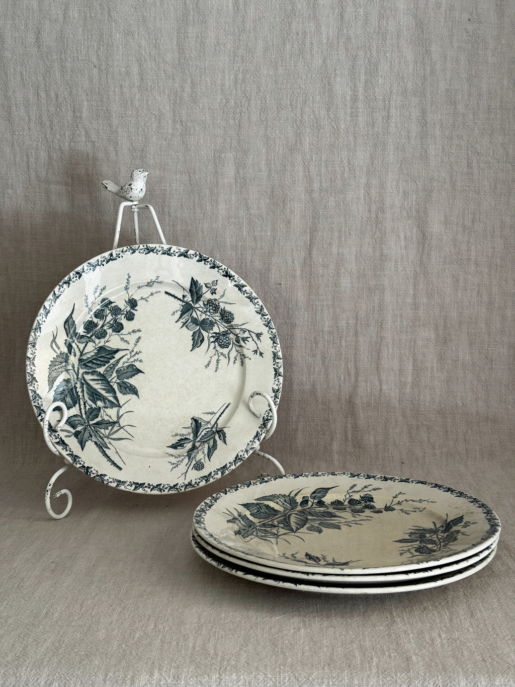 Four French green transferware dinner plates in a blackberry pattern, one upright on a white iron plate stand and three stacked on linen