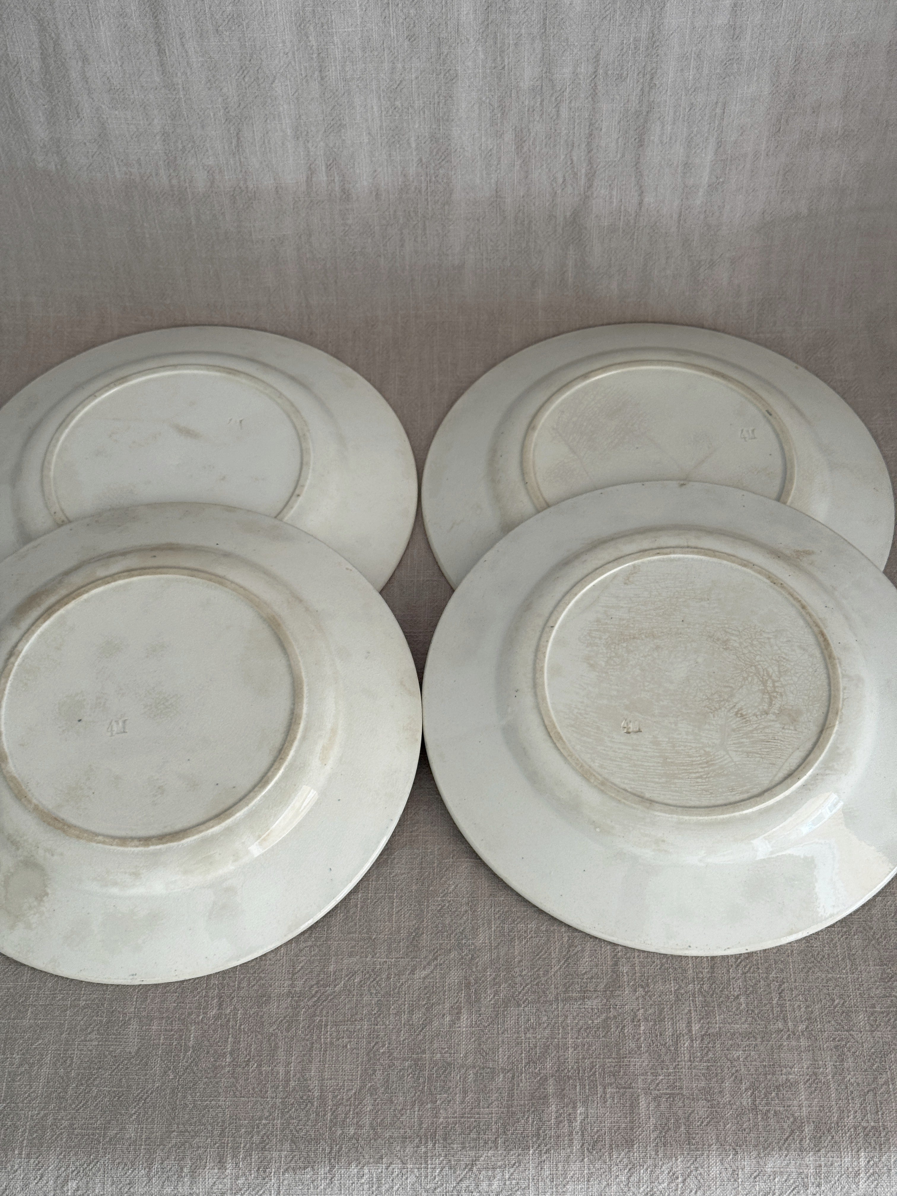 Undersides of four unmarked French luncheon plates laid out on linen, cream glaze with light age staining and impressed mould numbers